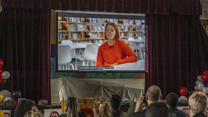 The documentary is projected onto a large screen in the gymnasium. In it, a woman is interviewed.
